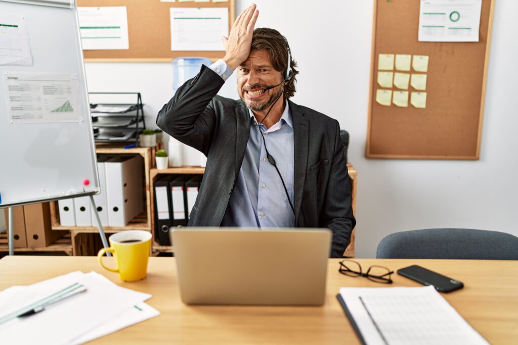 handsome middle age man wearing call center agent headset at the office surprised with hand on head for mistake, remember error. forgot, bad memory concept.